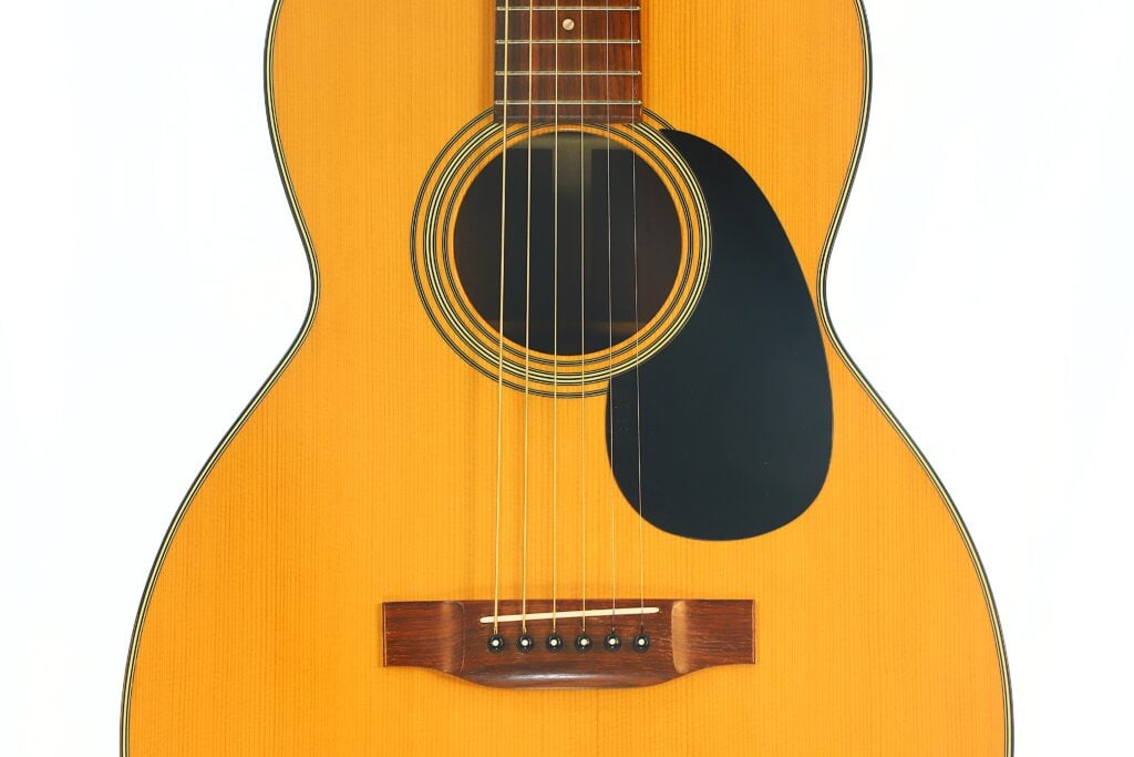 Nahaufnahme des Korpus der Martin 00-21 Akustikgitarre aus dem Jahr 1986, bei der die Saiten, das Schallloch, das Schlagbrett, der Holzsteg und die helle Decke vor weißem Hintergrund besonders hervorgehoben sind. Nahaufnahme des Korpus der Martin 00-21 Akustikgitarre aus dem Jahr 1986, bei der die Saiten, das Schallloch, das Schlagbrett, der Holzsteg und die helle Decke vor weißem Hintergrund besonders hervorgehoben sind.