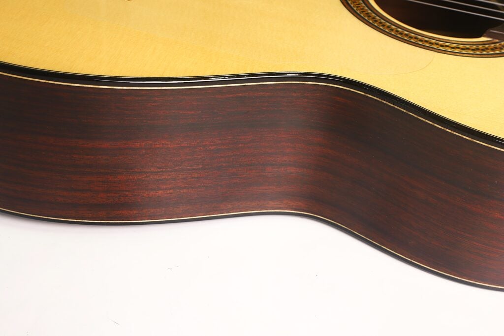 Nahaufnahme der Seite und Oberseite einer Lester DeVoe 2020 Negra Flamenco-Gitarre, die die glänzende Holzoberfläche, die eleganten Rundungen und den Rand des Schalllochs vor weißem Hintergrund hervorhebt. Nahaufnahme der Seite und Oberseite einer Lester DeVoe 2020 Negra Flamenco-Gitarre, die die glänzende Holzoberfläche, die eleganten Rundungen und den Rand des Schalllochs vor weißem Hintergrund hervorhebt.