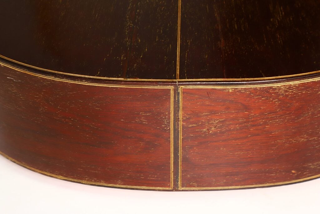 Nahaufnahme einer Holzoberfläche der „Klassische Gitarre“ von Andres Martin aus dem Jahr 1966, die sich durch einen dunklen oberen Bereich und einen rotbraunen unteren Bereich auszeichnet, die durch dünne goldene Linien voneinander getrennt sind, die ein rechteckiges Muster bilden. Nahaufnahme einer Holzoberfläche der „Klassische Gitarre“ von Andres Martin aus dem Jahr 1966, die sich durch einen dunklen oberen Bereich und einen rotbraunen unteren Bereich auszeichnet, die durch dünne goldene Linien voneinander getrennt sind, die ein rechteckiges Muster bilden.