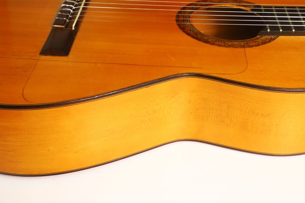 Nahaufnahme des Korpus einer Francisco-Barba-Flamenco-Gitarre aus dem Jahr 1966, auf der die Saiten, das Schallloch und die Holzoberfläche vor weißem Hintergrund zu sehen sind. Nahaufnahme des Korpus einer Francisco-Barba-Flamenco-Gitarre aus dem Jahr 1966, auf der die Saiten, das Schallloch und die Holzoberfläche vor weißem Hintergrund zu sehen sind.