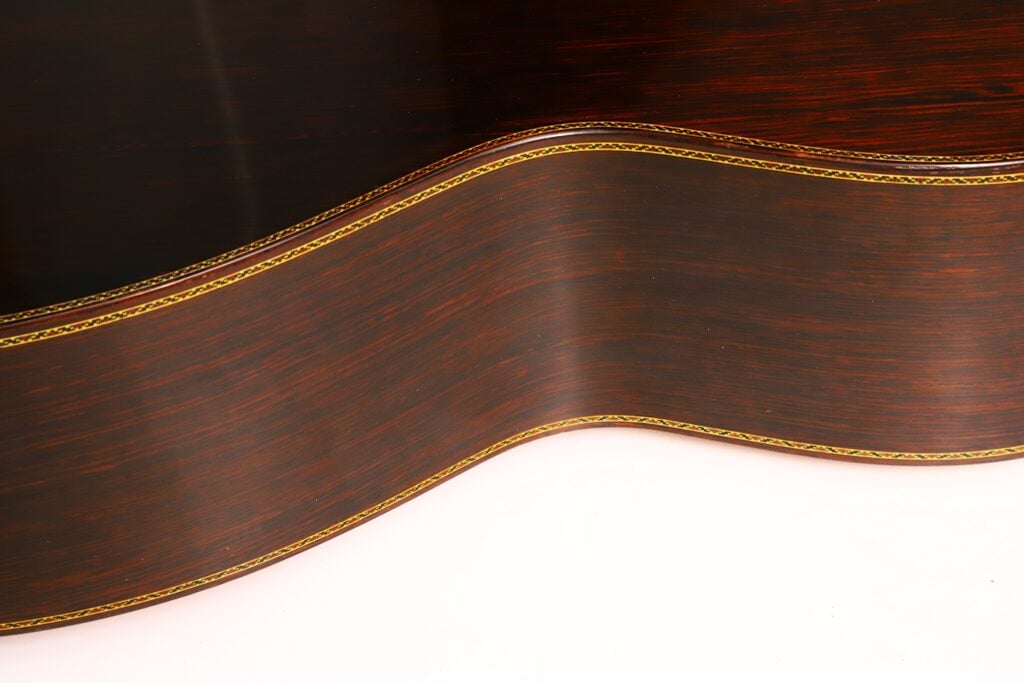 Nahaufnahme der dunklen Holzseite einer „Masaru Kohno Modell 50 1979“ Klassischen Gitarre, die ihre wellenförmige Krümmung und die dekorative gelbe Zierleiste vor einem hellen Hintergrund zeigt. Nahaufnahme der dunklen Holzseite einer „Masaru Kohno Modell 50 1979“ Klassischen Gitarre, die ihre wellenförmige Krümmung und die dekorative gelbe Zierleiste vor einem hellen Hintergrund zeigt.