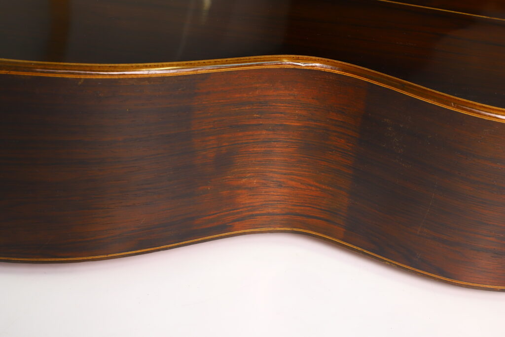 Nahaufnahme der Seite einer „Romantikgitarre“ von Louis Panormo aus dem Jahr 1827, die die Holzmaserung und die natürliche dunkelbraune Farbe mit glänzender Oberfläche und hellen Zierleisten zeigt. Nahaufnahme der Seite einer „Romantikgitarre“ von Louis Panormo aus dem Jahr 1827, die die Holzmaserung und die natürliche dunkelbraune Farbe mit glänzender Oberfläche und hellen Zierleisten zeigt.