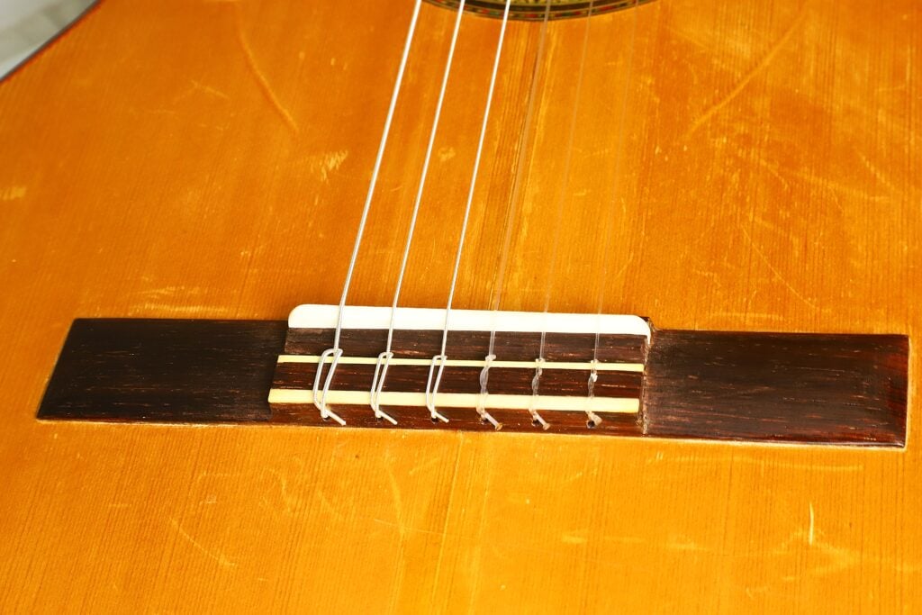 Eine detaillierte Nahaufnahme der „Klassische Gitarre“ von Andres Martin aus dem Jahr 1966 zeigt ihren Holzkorpus und hebt dabei den Steg, den Sattel und die Nylonsaiten vor dem Hintergrund einer warm polierten Oberfläche hervor. Eine detaillierte Nahaufnahme der „Klassische Gitarre“ von Andres Martin aus dem Jahr 1966 zeigt ihren Holzkorpus und hebt dabei den Steg, den Sattel und die Nylonsaiten vor dem Hintergrund einer warm polierten Oberfläche hervor.