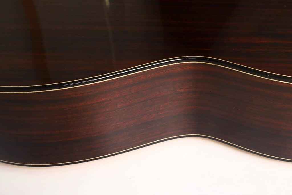 Nahaufnahme der Seite einer Lester DeVoe 2020 Negra Flamenco-Gitarre, die die elegante Korpuswölbung und die satte, dunkle Holzmaserung hervorhebt, die durch cremefarbene Zierleisten akzentuiert wird. Nahaufnahme der Seite einer Lester DeVoe 2020 Negra Flamenco-Gitarre, die die elegante Korpuswölbung und die satte, dunkle Holzmaserung hervorhebt, die durch cremefarbene Zierleisten akzentuiert wird.