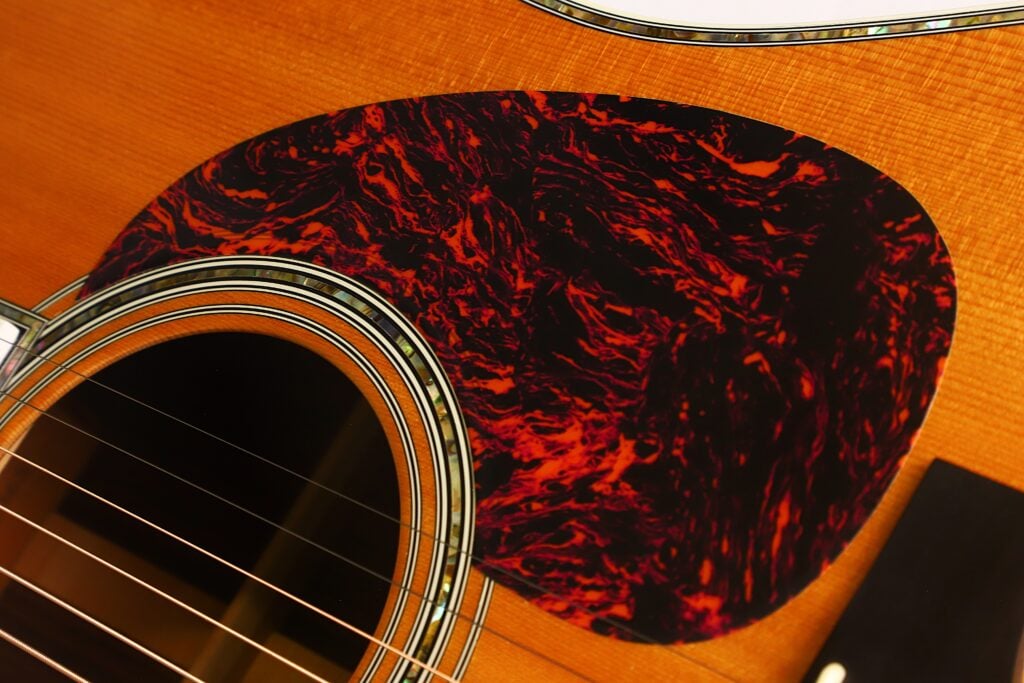 Nahaufnahme der Martin D-45 Akustikgitarre aus dem Jahr 2009, bei der die Saiten, ein Teil des Schalllochs und ein marmoriertes rot-schwarzes Schlagbrett auf dem Holzkorpus im Vordergrund stehen. Nahaufnahme der Martin D-45 Akustikgitarre aus dem Jahr 2009, bei der die Saiten, ein Teil des Schalllochs und ein marmoriertes rot-schwarzes Schlagbrett auf dem Holzkorpus im Vordergrund stehen.