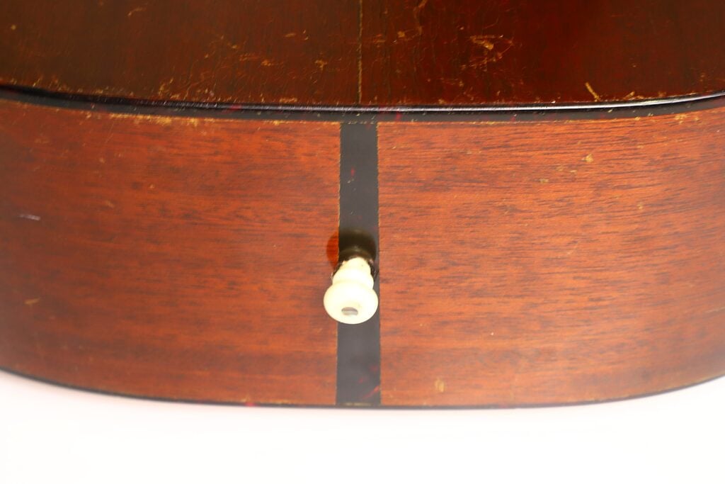 Nahaufnahme der Unterkante einer Martin 5-18 Akustikgitarre aus dem Jahr 1958, auf der in der Mitte ein weißer Endpin zu sehen ist, durch dessen Holzmaserung ein schwarzer vertikaler Streifen verläuft. Nahaufnahme der Unterkante einer Martin 5-18 Akustikgitarre aus dem Jahr 1958, auf der in der Mitte ein weißer Endpin zu sehen ist, durch dessen Holzmaserung ein schwarzer vertikaler Streifen verläuft.