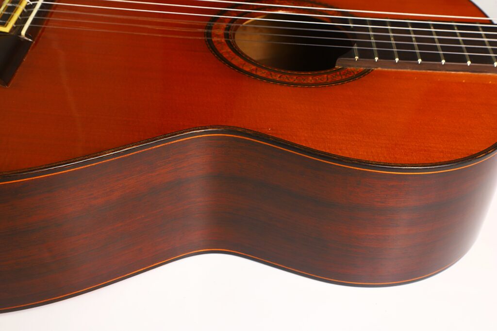 Nahaufnahme des Korpus einer Ramirez 1a 1985 Klassische Gitarre, bei der die Holzstruktur, die Saiten und das Schallloch hervorgehoben werden, wobei die Kanten vor einem weißen Hintergrund deutlich zu erkennen sind. Nahaufnahme des Korpus einer Ramirez 1a 1985 Klassische Gitarre, bei der die Holzstruktur, die Saiten und das Schallloch hervorgehoben werden, wobei die Kanten vor einem weißen Hintergrund deutlich zu erkennen sind.