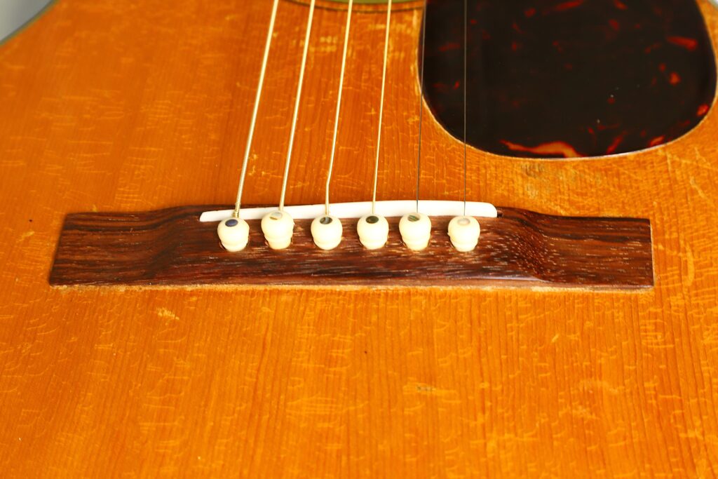 Nahaufnahme des Stegs einer Martin 5-18 Akustikgitarre aus dem Jahr 1958, auf dem sechs weiße Stifte zu sehen sind, mit denen die Saiten am Holzkorpus befestigt sind. In der oberen rechten Ecke ist teilweise das Schlagbrett zu erkennen. Nahaufnahme des Stegs einer Martin 5-18 Akustikgitarre aus dem Jahr 1958, auf dem sechs weiße Stifte zu sehen sind, mit denen die Saiten am Holzkorpus befestigt sind. In der oberen rechten Ecke ist teilweise das Schlagbrett zu erkennen.