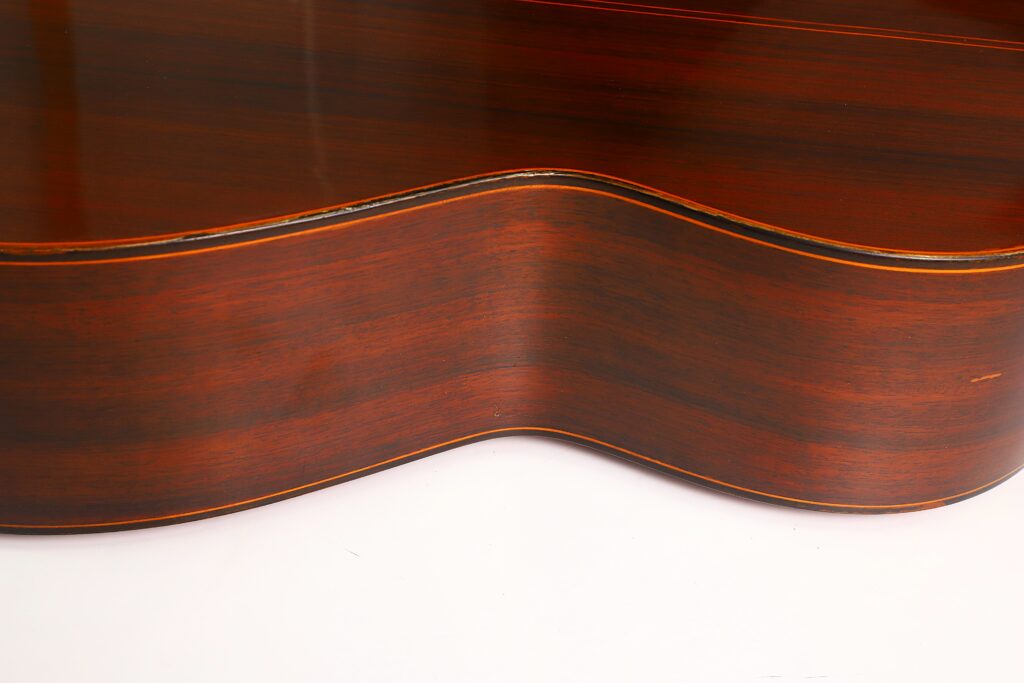 Nahaufnahme der geschwungenen Holzseite der Ramirez 1a 1985 Klassische Gitarre, die die glatte braune Holzmaserung und die schwarzen Einfassungen vor weißem Hintergrund hervorhebt. Nahaufnahme der geschwungenen Holzseite der Ramirez 1a 1985 Klassische Gitarre, die die glatte braune Holzmaserung und die schwarzen Einfassungen vor weißem Hintergrund hervorhebt.