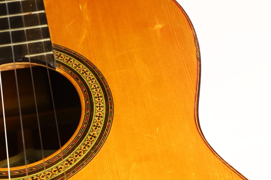 Nahaufnahme des Korpus einer „Klassische Gitarre“ von Andres Martin aus dem Jahr 1966, bei der das Schallloch, die Zierrosette, die Saiten und ein Teil des Griffbretts vor weißem Hintergrund hervorgehoben sind. Nahaufnahme des Korpus einer „Klassische Gitarre“ von Andres Martin aus dem Jahr 1966, bei der das Schallloch, die Zierrosette, die Saiten und ein Teil des Griffbretts vor weißem Hintergrund hervorgehoben sind.