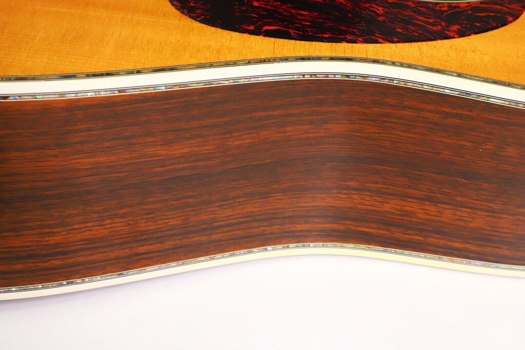 Nahaufnahme der Seite einer Martin D-45 Akustikgitarre aus dem Jahr 2009, die den geschwungenen Holzkorpus mit glänzender Lackierung, die feine Holzmaserung und die kunstvolle Zierleiste an den Kanten hervorhebt. Nahaufnahme der Seite einer Martin D-45 Akustikgitarre aus dem Jahr 2009, die den geschwungenen Holzkorpus mit glänzender Lackierung, die feine Holzmaserung und die kunstvolle Zierleiste an den Kanten hervorhebt.