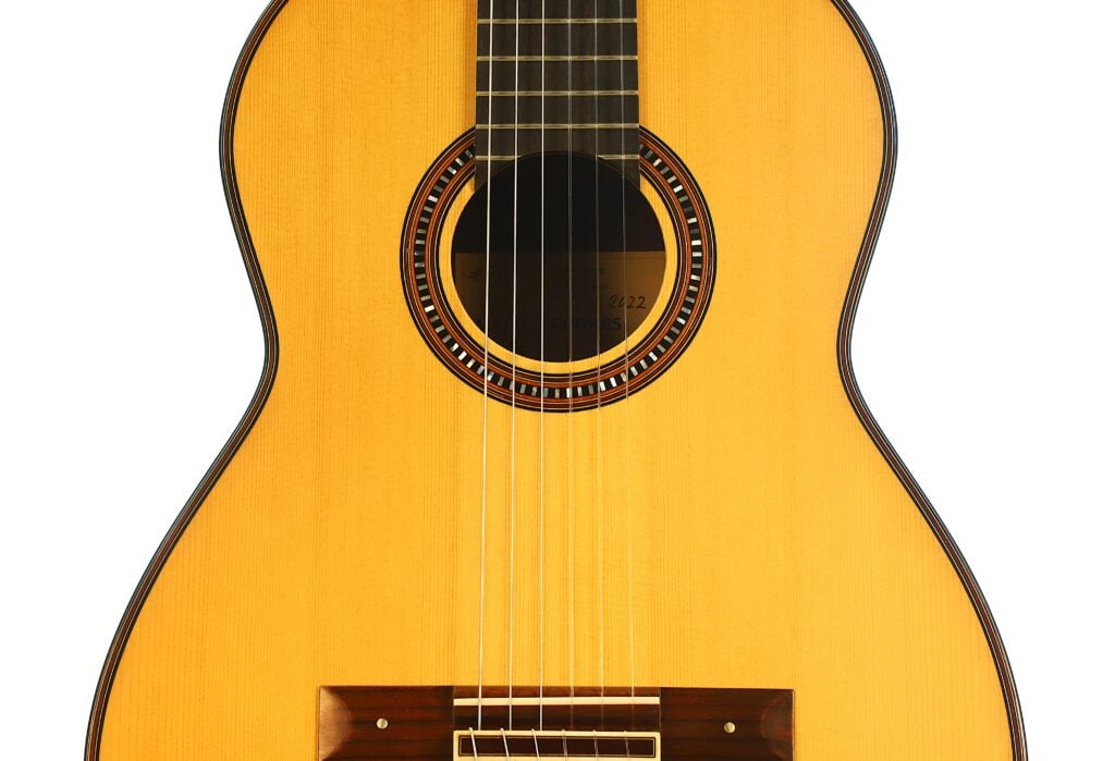 Nahaufnahme der Vorderseite einer „Hohlkehle Klassische Gitarre“ von Matthias Hartig aus dem Jahr 2022, die sechs Saiten, das Schallloch mit dekorativer Rosette und einen Teil des Holzstegs vor weißem Hintergrund zeigt. Nahaufnahme der Vorderseite einer „Hohlkehle Klassische Gitarre“ von Matthias Hartig aus dem Jahr 2022, die sechs Saiten, das Schallloch mit dekorativer Rosette und einen Teil des Holzstegs vor weißem Hintergrund zeigt.