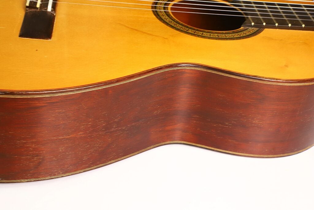 Nahaufnahme des Korpus einer „Klassische Gitarre“ von Andres Martin aus dem Jahr 1966, die die Maserung der Zargen und der Decke hervorhebt; vor weißem Hintergrund sind die Saiten und ein Teil des Schalllochs zu sehen. Nahaufnahme des Korpus einer „Klassische Gitarre“ von Andres Martin aus dem Jahr 1966, die die Maserung der Zargen und der Decke hervorhebt; vor weißem Hintergrund sind die Saiten und ein Teil des Schalllochs zu sehen.