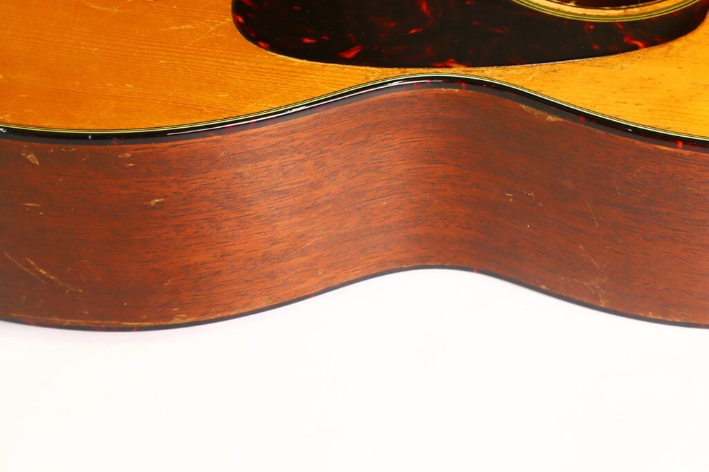 Nahaufnahme der Seite einer Martin 5-18 Akustikgitarre aus dem Jahr 1958, bei der die Maserung des Holzes, die geschwungene Form und ein Teil der Decke der Gitarre vor einem weißen Hintergrund hervorgehoben werden. Nahaufnahme der Seite einer Martin 5-18 Akustikgitarre aus dem Jahr 1958, bei der die Maserung des Holzes, die geschwungene Form und ein Teil der Decke der Gitarre vor einem weißen Hintergrund hervorgehoben werden.