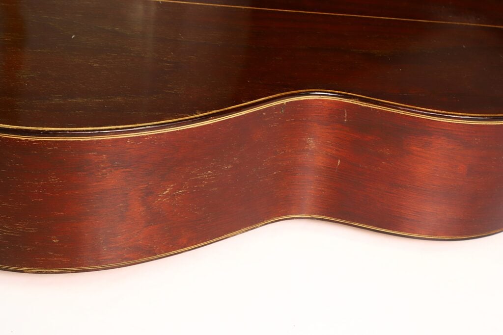 Seitenansicht der „Andres Martin 1966 Klassische Gitarre“, die den geschwungenen Korpus, die glänzende rotbraune Lackierung mit markanter Holzmaserung und die detailreiche, feine Veredelung hervorhebt. Seitenansicht der „Andres Martin 1966 Klassische Gitarre“, die den geschwungenen Korpus, die glänzende rotbraune Lackierung mit markanter Holzmaserung und die detailreiche, feine Veredelung hervorhebt.