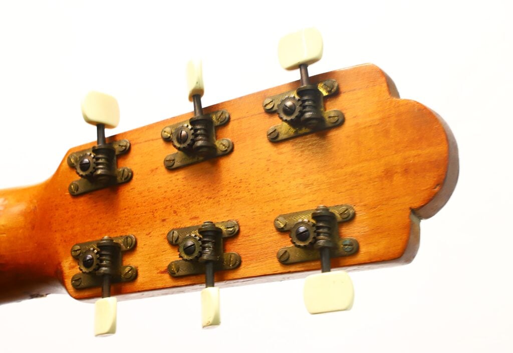 Nahaufnahme der Rückseite des Kopfes einer „Klassische Gitarre“ von Salvador Ibanez aus dem Jahr 1920, auf der sechs Stimmwirbel mit weißen Knöpfen und Metallbeschlägen vor einem weißen Hintergrund zu sehen sind. Nahaufnahme der Rückseite des Kopfes einer „Klassische Gitarre“ von Salvador Ibanez aus dem Jahr 1920, auf der sechs Stimmwirbel mit weißen Knöpfen und Metallbeschlägen vor einem weißen Hintergrund zu sehen sind.