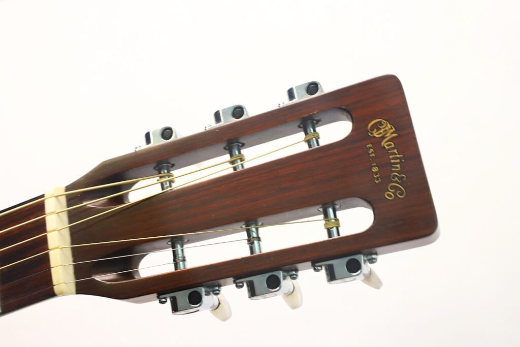 Nahaufnahme des Kopfes einer Martin 00-21 Akustikgitarre aus dem Jahr 1986 mit sechs Stimmwirbeln und goldenem Logo, auf der die Saiten und die Holzmaserung vor einem schlichten weißen Hintergrund zu sehen sind. Nahaufnahme des Kopfes einer Martin 00-21 Akustikgitarre aus dem Jahr 1986 mit sechs Stimmwirbeln und goldenem Logo, auf der die Saiten und die Holzmaserung vor einem schlichten weißen Hintergrund zu sehen sind.