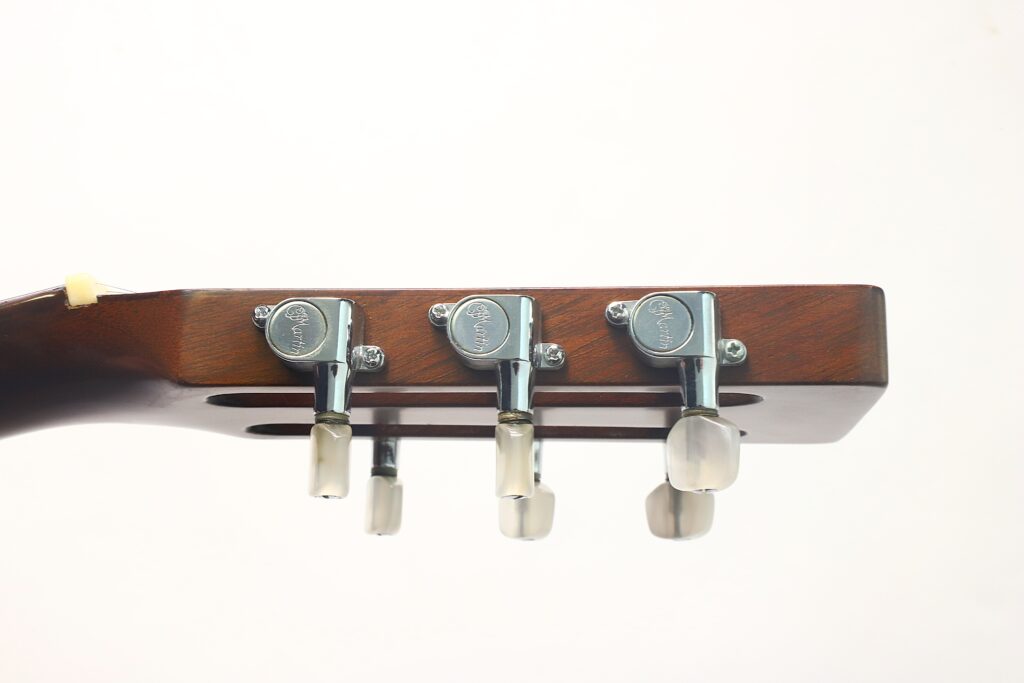 Nahaufnahme der Seitenansicht des Holzkopfes der Martin 00-21 Akustikgitarre aus dem Jahr 1986, mit drei silbernen Stimmwirbeln mit cremefarbenen Stimmknöpfen vor einem schlichten weißen Hintergrund. Nahaufnahme der Seitenansicht des Holzkopfes der Martin 00-21 Akustikgitarre aus dem Jahr 1986, mit drei silbernen Stimmwirbeln mit cremefarbenen Stimmknöpfen vor einem schlichten weißen Hintergrund.