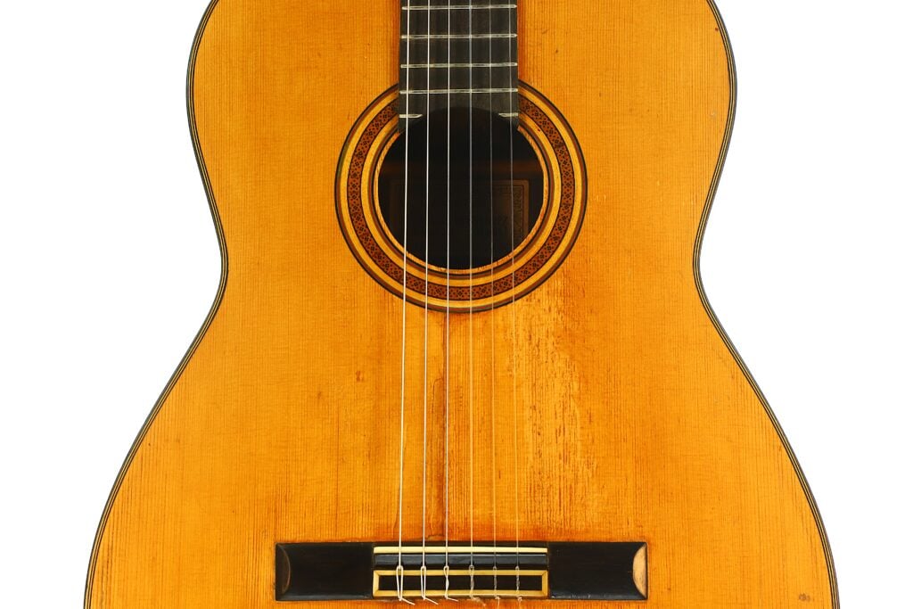 Eine Nahaufnahme des Korpus der „Angel Alonso 1936 Klassische Gitarre“, auf der die sechs Saiten, der Holzsteg, die aufwendig gearbeitete Rosette um das Schallloch sowie die naturbelassene Holzoberfläche zu sehen sind. Eine Nahaufnahme des Korpus der „Angel Alonso 1936 Klassische Gitarre“, auf der die sechs Saiten, der Holzsteg, die aufwendig gearbeitete Rosette um das Schallloch sowie die naturbelassene Holzoberfläche zu sehen sind.