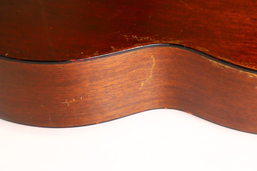 Nahaufnahme der Seite einer Martin 5-18 Akustikgitarre aus dem Jahr 1958, die die Holzmaserung, kleinere Kratzer und einige Gebrauchsspuren auf der Hochglanzoberfläche zeigt. Nahaufnahme der Seite einer Martin 5-18 Akustikgitarre aus dem Jahr 1958, die die Holzmaserung, kleinere Kratzer und einige Gebrauchsspuren auf der Hochglanzoberfläche zeigt.