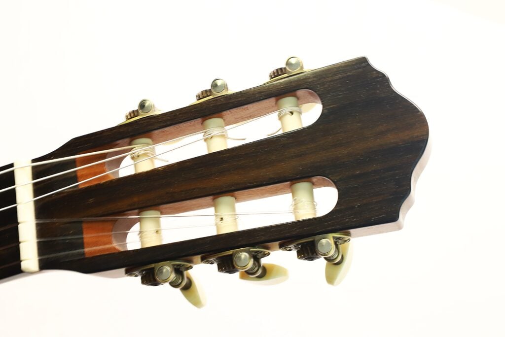 Nahaufnahme des Kopfes der „Matthias Hartig 2022 Hohlkehle Klassische Gitarre“ mit Stimmwirbeln, Saiten und einer dunklen Holzoberfläche vor einem schlichten weißen Hintergrund. Nahaufnahme des Kopfes der „Matthias Hartig 2022 Hohlkehle Klassische Gitarre“ mit Stimmwirbeln, Saiten und einer dunklen Holzoberfläche vor einem schlichten weißen Hintergrund.