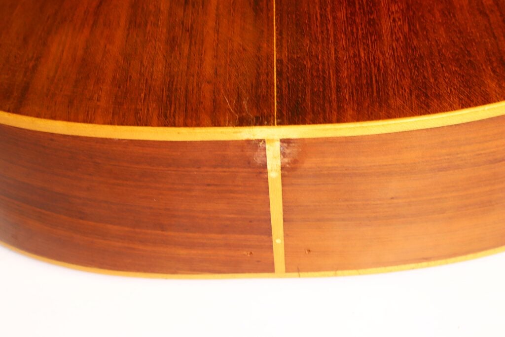 Nahaufnahme der Seite und Rückseite der „Salvador Ibanez 1920 Klassische Gitarre“, die die Maserung des Holzes, die helle Einfassung und eine vertikale Fuge an der Verbindungsstelle zweier Holzstücke hervorhebt. Nahaufnahme der Seite und Rückseite der „Salvador Ibanez 1920 Klassische Gitarre“, die die Maserung des Holzes, die helle Einfassung und eine vertikale Fuge an der Verbindungsstelle zweier Holzstücke hervorhebt.