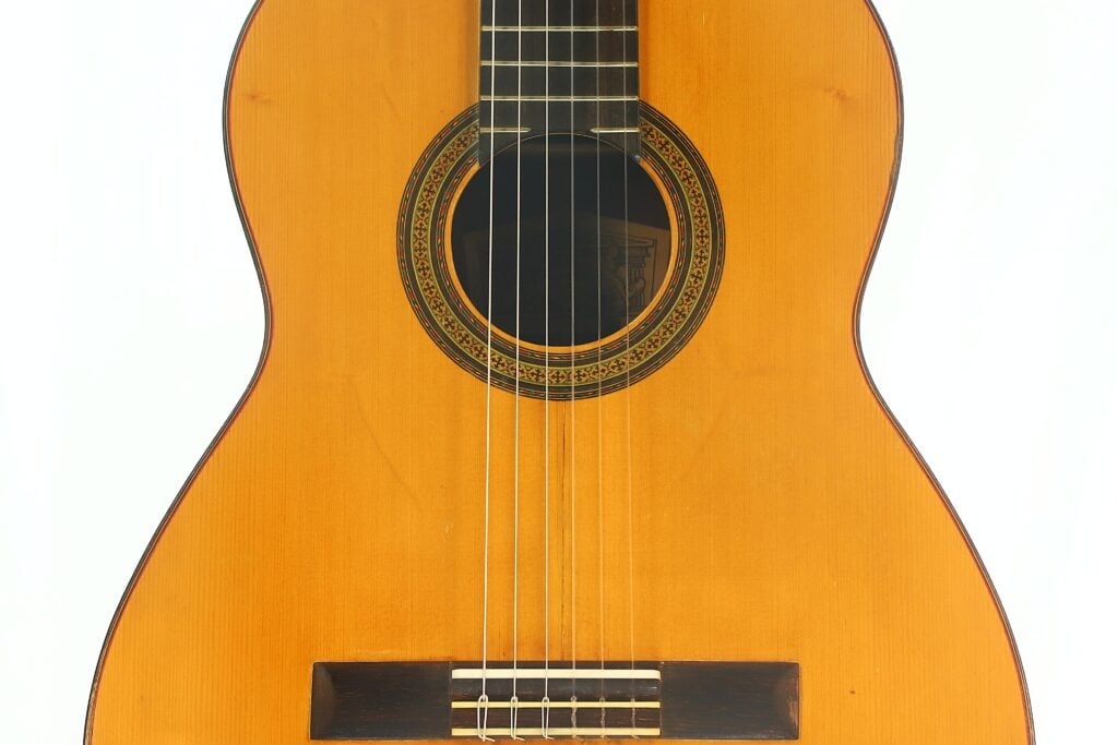 Eine detaillierte Nahaufnahme der „Klassische Gitarre“ von Andres Martin aus dem Jahr 1966 zeigt den Korpus mit dem durch eine Zierrosette verzierten Schallloch, den sichtbaren Saiten und einem Ausschnitt des Griffbretts vor weißem Hintergrund. Eine detaillierte Nahaufnahme der „Klassische Gitarre“ von Andres Martin aus dem Jahr 1966 zeigt den Korpus mit dem durch eine Zierrosette verzierten Schallloch, den sichtbaren Saiten und einem Ausschnitt des Griffbretts vor weißem Hintergrund.