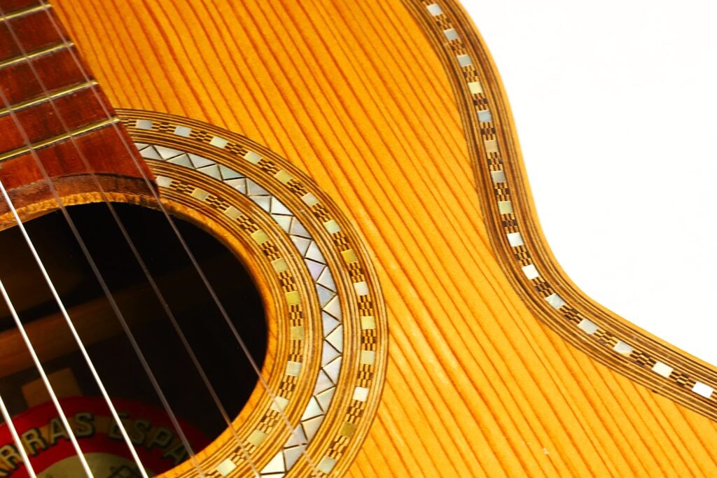 Nahaufnahme des Korpus einer „Klassische Gitarre“ von Salvador Ibanez aus dem Jahr 1920, bei der das Schallloch mit seiner dekorativen Rosette, die detaillierte Holzmaserung sowie ein Teil der Saiten auf der linken Seite vor einem weißen Hintergrund hervorgehoben werden. Nahaufnahme des Korpus einer „Klassische Gitarre“ von Salvador Ibanez aus dem Jahr 1920, bei der das Schallloch mit seiner dekorativen Rosette, die detaillierte Holzmaserung sowie ein Teil der Saiten auf der linken Seite vor einem weißen Hintergrund hervorgehoben werden.
