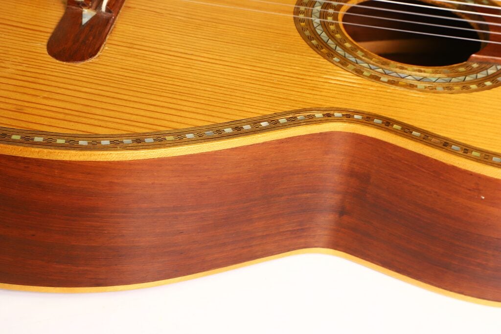 Nahaufnahme des Korpus der „Salvador Ibanez 1920 Klassische Gitarre“, die die Holzmaserung, die glänzende Lackierung, die dekorative Rosette um das Schallloch sowie mehrere Saiten hervorhebt. Die Gitarre zeichnet sich durch kontrastierende helle und dunkle Holztöne aus. Nahaufnahme des Korpus der „Salvador Ibanez 1920 Klassische Gitarre“, die die Holzmaserung, die glänzende Lackierung, die dekorative Rosette um das Schallloch sowie mehrere Saiten hervorhebt. Die Gitarre zeichnet sich durch kontrastierende helle und dunkle Holztöne aus.
