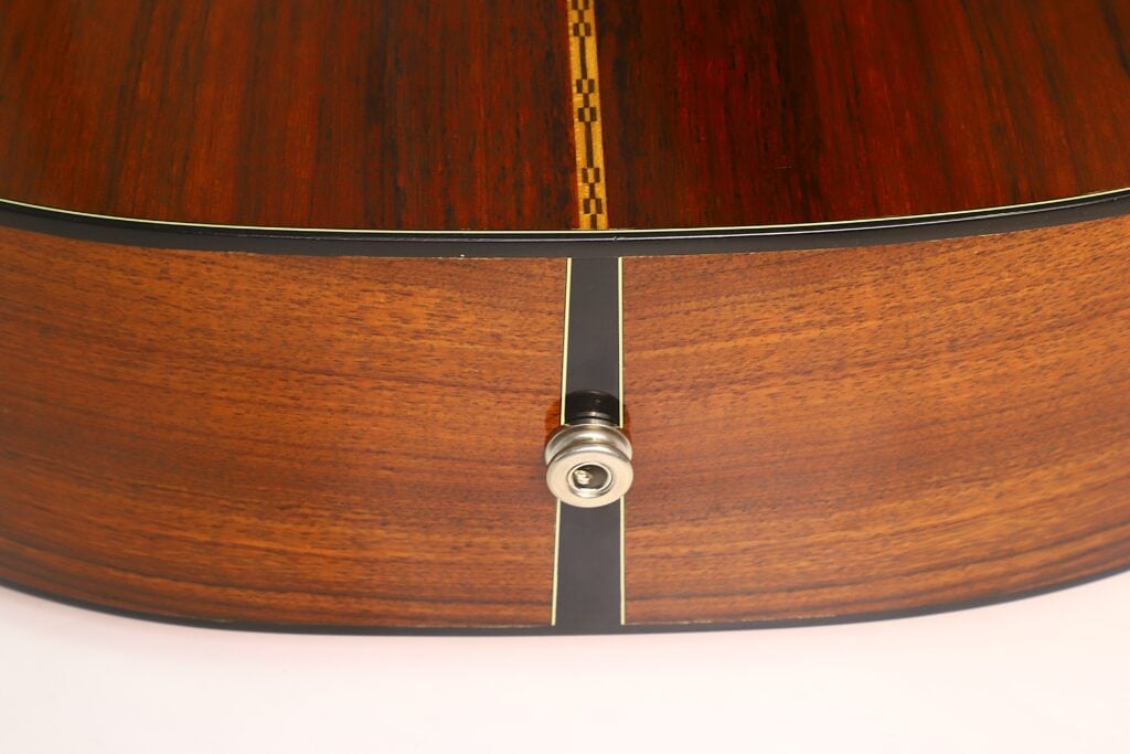 Nahaufnahme der Unterseite einer Martin 00-21 Akustikgitarre aus dem Jahr 1986, auf der ein am Holz befestigter Metall-Gurtknopf zu sehen ist, mit sichtbarer Maserung und einer dekorativen Einlage, die entlang der Mittellinie verläuft. Nahaufnahme der Unterseite einer Martin 00-21 Akustikgitarre aus dem Jahr 1986, auf der ein am Holz befestigter Metall-Gurtknopf zu sehen ist, mit sichtbarer Maserung und einer dekorativen Einlage, die entlang der Mittellinie verläuft.