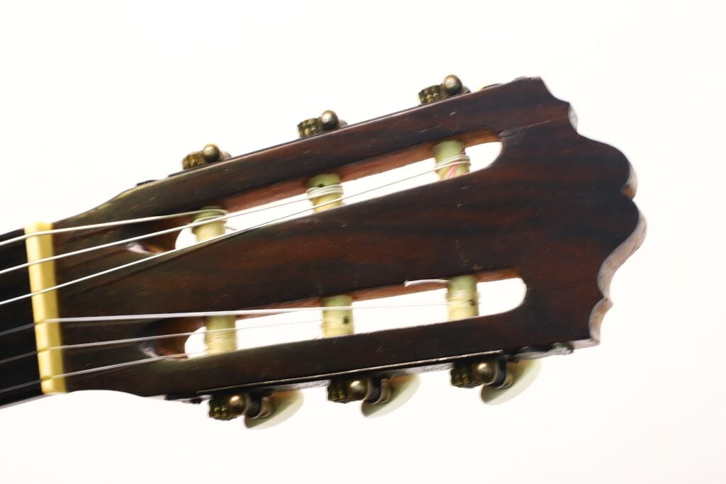 Eine Nahaufnahme des Kopfes der „Angel Alonso 1936 Santos Hernan“ Klassische Gitarre, die die Stimmwirbel, die detaillierten Saitenwicklungen und die kräftige Holzmaserung vor weißem Hintergrund zeigt. Eine Nahaufnahme des Kopfes der „Angel Alonso 1936 Santos Hernan“ Klassische Gitarre, die die Stimmwirbel, die detaillierten Saitenwicklungen und die kräftige Holzmaserung vor weißem Hintergrund zeigt.