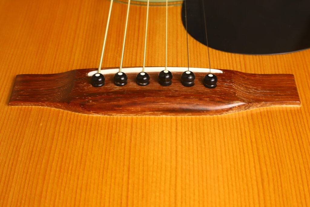 Nahaufnahme des Stegs einer Martin 00-21 Akustikgitarre aus dem Jahr 1986, auf der sechs Saiten zu sehen sind, die mit schwarzen Stegstiften auf einer polierten Holzoberfläche befestigt sind. Nahaufnahme des Stegs einer Martin 00-21 Akustikgitarre aus dem Jahr 1986, auf der sechs Saiten zu sehen sind, die mit schwarzen Stegstiften auf einer polierten Holzoberfläche befestigt sind.