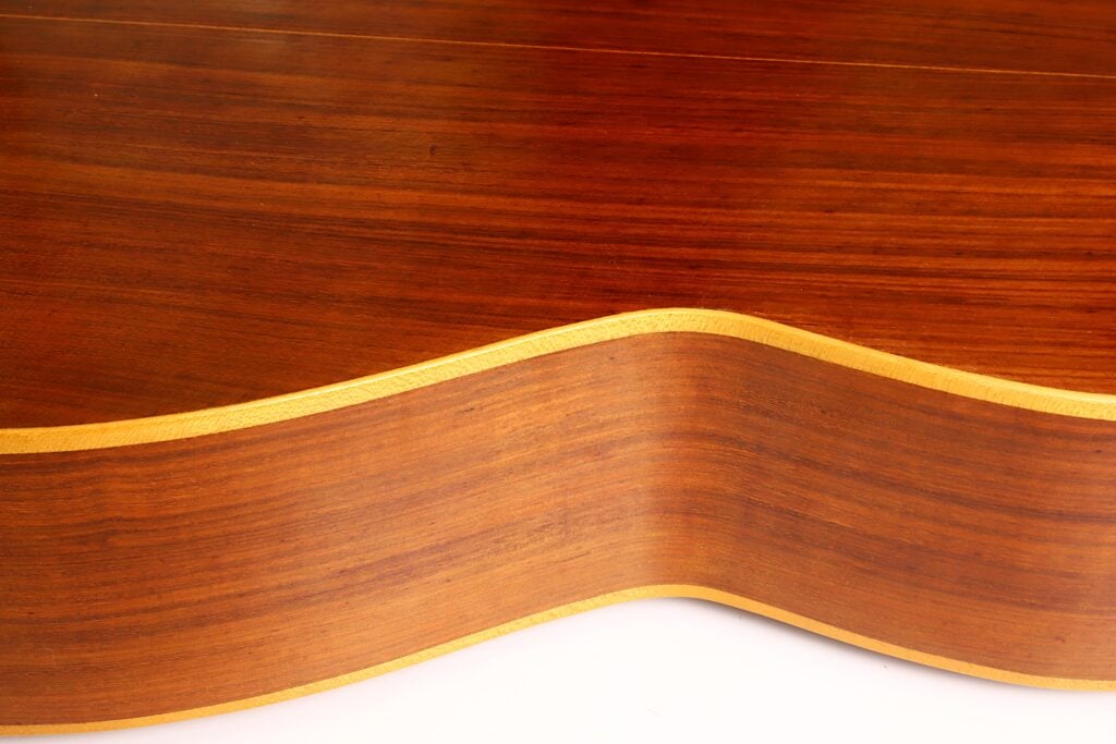 Nahaufnahme der Seite der „Salvador Ibanez 1920 Klassische Gitarre“, die die glatte, geschwungene Holzmaserung mit einem helleren Rand vor weißem Hintergrund hervorhebt. Nahaufnahme der Seite der „Salvador Ibanez 1920 Klassische Gitarre“, die die glatte, geschwungene Holzmaserung mit einem helleren Rand vor weißem Hintergrund hervorhebt.