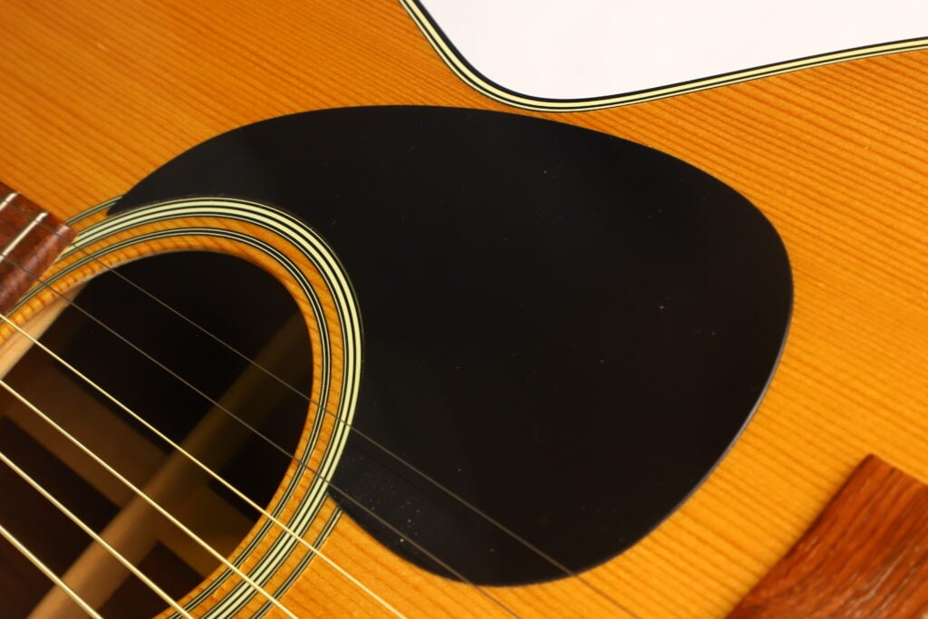 Nahaufnahme einer Martin 00-21 Akustikgitarre aus dem Jahr 1986, auf der ein Teil des Schalllochs, die Saiten, das schwarze Schlagbrett und die helle Holzmaserung des Gitarrenkorpus zu sehen sind. Nahaufnahme einer Martin 00-21 Akustikgitarre aus dem Jahr 1986, auf der ein Teil des Schalllochs, die Saiten, das schwarze Schlagbrett und die helle Holzmaserung des Gitarrenkorpus zu sehen sind.
