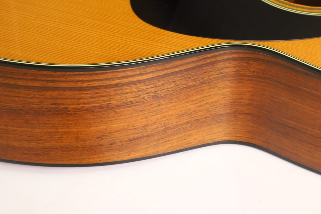 Nahaufnahme der Seite einer Martin 00-21 Akustikgitarre aus dem Jahr 1986, die die Holzmaserung und einen Teil der glänzenden Decke mit schwarzem Schlagbrett hervorhebt. Nahaufnahme der Seite einer Martin 00-21 Akustikgitarre aus dem Jahr 1986, die die Holzmaserung und einen Teil der glänzenden Decke mit schwarzem Schlagbrett hervorhebt.