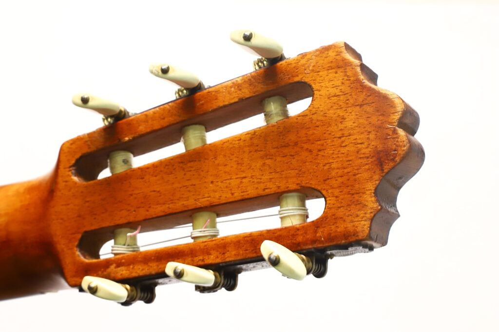 Nahaufnahme des Kopfstücks der „Angel Alonso 1936 Santos Hernan“ Klassische Gitarre mit sechs Stimmwirbeln und einer Holzoberfläche vor einem schlichten weißen Hintergrund. Nahaufnahme des Kopfstücks der „Angel Alonso 1936 Santos Hernan“ Klassische Gitarre mit sechs Stimmwirbeln und einer Holzoberfläche vor einem schlichten weißen Hintergrund.