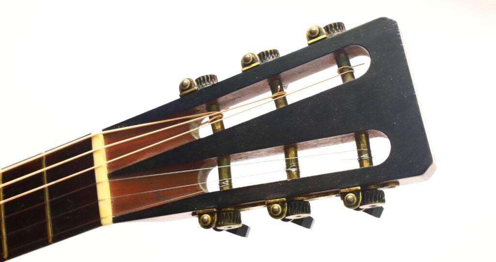 Nahaufnahme des Kopfes der James Ashborn 1850 Parlor-Akustikgitarre mit geschlitztem Design, offenen Mechaniken im Vintage-Stil und sechs Stimmwirbeln, abgebildet mit Metallsaiten vor weißem Hintergrund. Nahaufnahme des Kopfes der James Ashborn 1850 Parlor-Akustikgitarre mit geschlitztem Design, offenen Mechaniken im Vintage-Stil und sechs Stimmwirbeln, abgebildet mit Metallsaiten vor weißem Hintergrund.