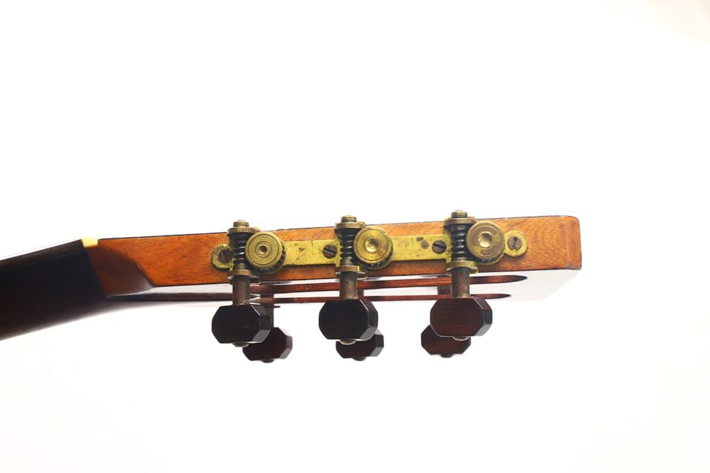 Nahaufnahme des Kopfstücks einer James Ashborn Parlor-Akustikgitarre aus dem Jahr 1850, mit drei dunklen Stimmwirbeln mit Messingbeschlägen vor einem schlichten weißen Hintergrund. Nahaufnahme des Kopfstücks einer James Ashborn Parlor-Akustikgitarre aus dem Jahr 1850, mit drei dunklen Stimmwirbeln mit Messingbeschlägen vor einem schlichten weißen Hintergrund.