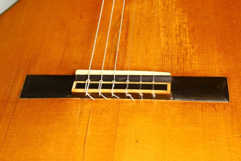 Nahaufnahme der Brücke und der Saiten der klassischen Gitarre „Angel Alonso 1936 Santos Hernan“ auf ihrem Holzkorpus, die die feinen Details der Bauweise dieser klassischen Gitarre hervorhebt. Nahaufnahme der Brücke und der Saiten der klassischen Gitarre „Angel Alonso 1936 Santos Hernan“ auf ihrem Holzkorpus, die die feinen Details der Bauweise dieser klassischen Gitarre hervorhebt.