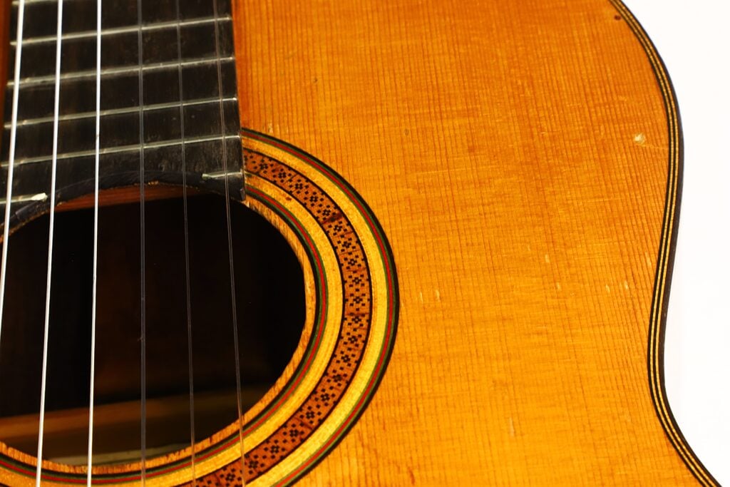 Nahaufnahme einer „Angel Alonso 1936 Santos Hernan“-Klassikgitarre, bei der die Saiten, das Schallloch und ein Teil der dekorativen Rosette auf dem Holzfinish besonders hervorgehoben sind. Nahaufnahme einer „Angel Alonso 1936 Santos Hernan“-Klassikgitarre, bei der die Saiten, das Schallloch und ein Teil der dekorativen Rosette auf dem Holzfinish besonders hervorgehoben sind.
