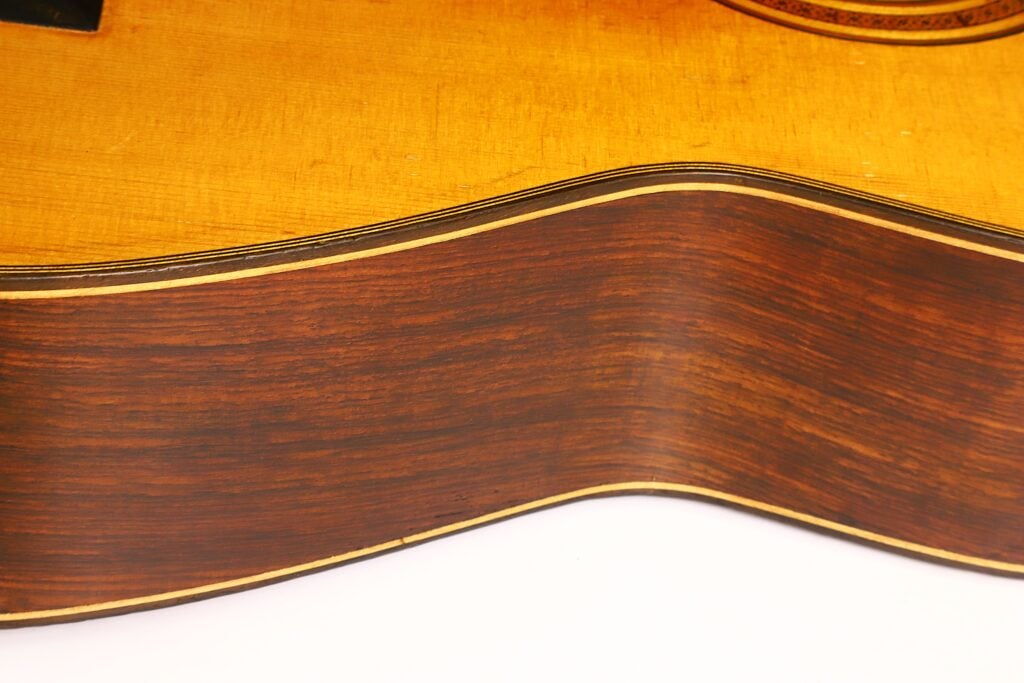 Nahaufnahme der Seite der „Angel Alonso 1936 Klassische Gitarre – Santos Hernandez“, die die Holzmaserung und die elegant geschwungene Kante hervorhebt, mit einem gelblichen oberen Bereich und einer dunkelbraunen Seite, die durch einen schmalen cremefarbenen Streifen voneinander getrennt sind. Nahaufnahme der Seite der „Angel Alonso 1936 Klassische Gitarre – Santos Hernandez“, die die Holzmaserung und die elegant geschwungene Kante hervorhebt, mit einem gelblichen oberen Bereich und einer dunkelbraunen Seite, die durch einen schmalen cremefarbenen Streifen voneinander getrennt sind.
