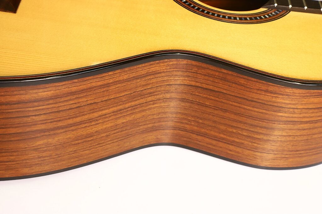 Nahaufnahme der Seite und eines Teils der Oberseite der „Matthias Hartig 2022 Hohlkehle Klassische Gitarre“, die die Holzmaserung und die glatte Oberfläche hervorhebt, wobei der Fokus auf den eleganten Rundungen und dem strukturierten Korpus des Instruments liegt. Nahaufnahme der Seite und eines Teils der Oberseite der „Matthias Hartig 2022 Hohlkehle Klassische Gitarre“, die die Holzmaserung und die glatte Oberfläche hervorhebt, wobei der Fokus auf den eleganten Rundungen und dem strukturierten Korpus des Instruments liegt.