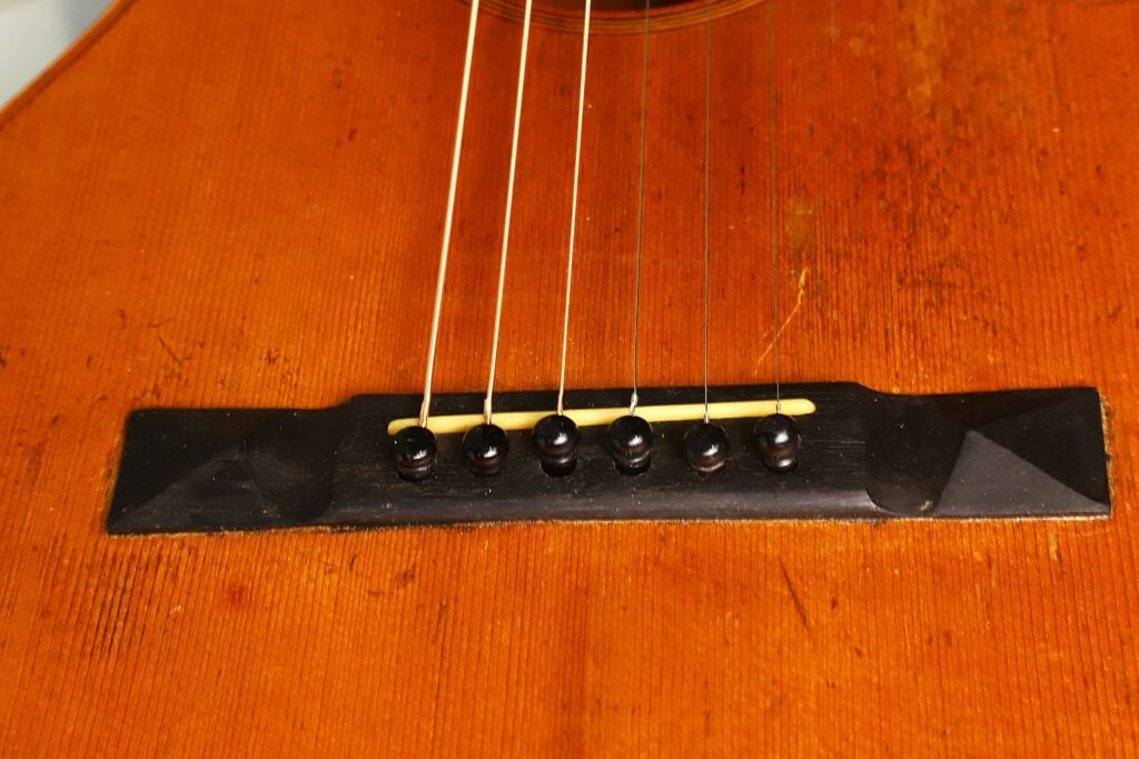 Nahaufnahme des Stegs der James Ashborn 1850 Parlor Akustikgitarre, auf der sechs Saiten zu sehen sind, die mit schwarzen Stegstiften auf einem warmbraunen Holzkorpus befestigt sind. Nahaufnahme des Stegs der James Ashborn 1850 Parlor Akustikgitarre, auf der sechs Saiten zu sehen sind, die mit schwarzen Stegstiften auf einem warmbraunen Holzkorpus befestigt sind.