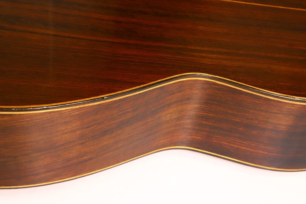 Nahaufnahme der Seite der „Angel Alonso 1936 Klassische Gitarre – Santos Hernandez“, die die elegant geschwungene Holzmaserung und die dekorative Kantenverzierung zeigt. Der weiße Hintergrund bringt die satte braune Lackierung der Gitarre besonders gut zur Geltung. Nahaufnahme der Seite der „Angel Alonso 1936 Klassische Gitarre – Santos Hernandez“, die die elegant geschwungene Holzmaserung und die dekorative Kantenverzierung zeigt. Der weiße Hintergrund bringt die satte braune Lackierung der Gitarre besonders gut zur Geltung.