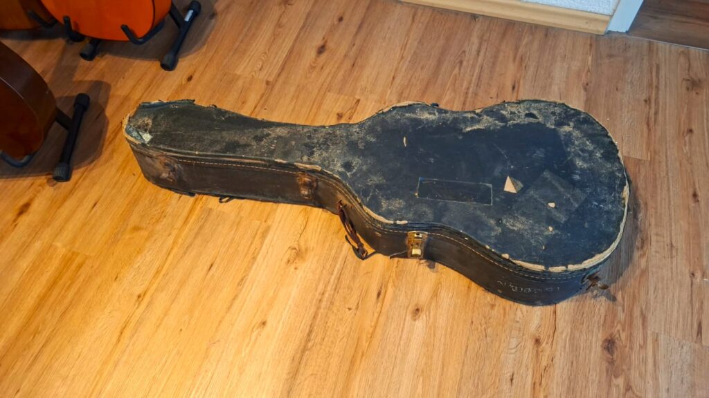 Ein abgenutzter, schwarzer Hartschalenkoffer für eine Martin D-28 1941 Pre-War Akustikgitarre mit abblätternder Lackierung und beschädigten Kanten liegt geschlossen auf einem Holzboden. Ein abgenutzter, schwarzer Hartschalenkoffer für eine Martin D-28 1941 Pre-War Akustikgitarre mit abblätternder Lackierung und beschädigten Kanten liegt geschlossen auf einem Holzboden.