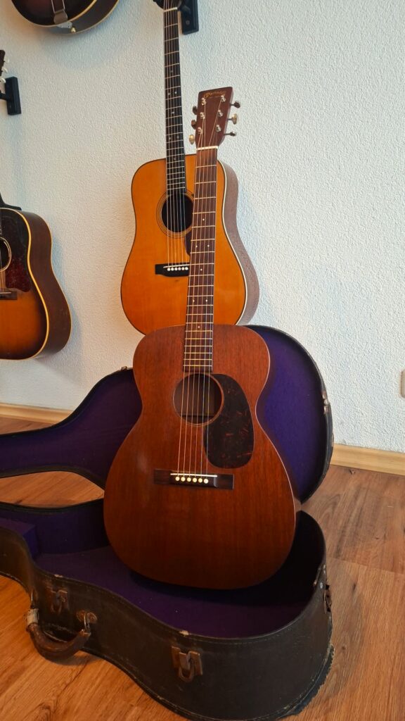 Zu sehen sind zwei Martin 00-17 Akustikgitarren aus dem Jahr 1941; eine steht aufrecht in einem schwarzen Koffer mit violettem Innenfutter, während die andere im Hintergrund an der Wand hängt. Beide Gitarren haben eine Holzoberfläche und sechs Saiten. Zu sehen sind zwei Martin 00-17 Akustikgitarren aus dem Jahr 1941; eine steht aufrecht in einem schwarzen Koffer mit violettem Innenfutter, während die andere im Hintergrund an der Wand hängt. Beide Gitarren haben eine Holzoberfläche und sechs Saiten.