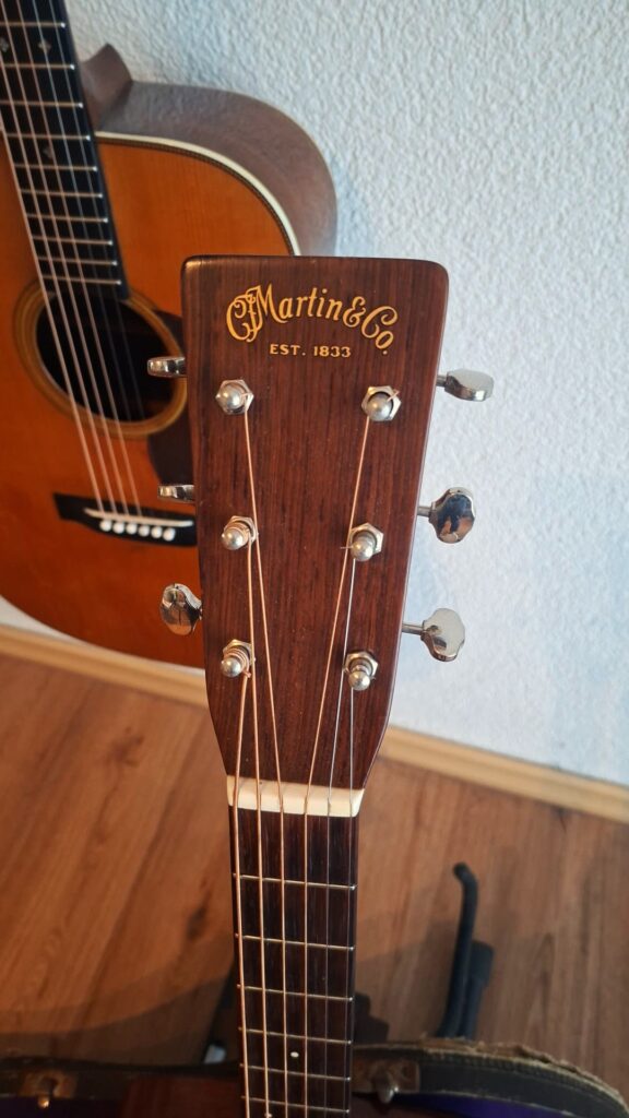 Nahaufnahme des Kopfes einer Martin 00-17 Akustikgitarre aus dem Jahr 1941 mit Stimmwirbeln, auf denen das Markenlogo zu sehen ist. Im Hintergrund hängt eine weitere Gitarre vor einer strukturierten weißen Wand auf einem Holzboden. Nahaufnahme des Kopfes einer Martin 00-17 Akustikgitarre aus dem Jahr 1941 mit Stimmwirbeln, auf denen das Markenlogo zu sehen ist. Im Hintergrund hängt eine weitere Gitarre vor einer strukturierten weißen Wand auf einem Holzboden.