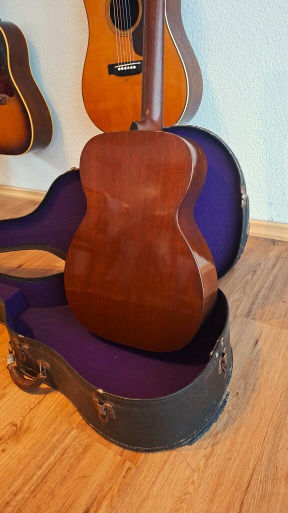 Eine Martin 00-17 Akustikgitarre aus dem Jahr 1941, deren Rückseite nach vorne zeigt, steht in einem offenen, mit lila gefütterten schwarzen Koffer, der auf einem Holzboden steht. Im Hintergrund hängen weitere Gitarren an einer hellen Wand. Eine Martin 00-17 Akustikgitarre aus dem Jahr 1941, deren Rückseite nach vorne zeigt, steht in einem offenen, mit lila gefütterten schwarzen Koffer, der auf einem Holzboden steht. Im Hintergrund hängen weitere Gitarren an einer hellen Wand.