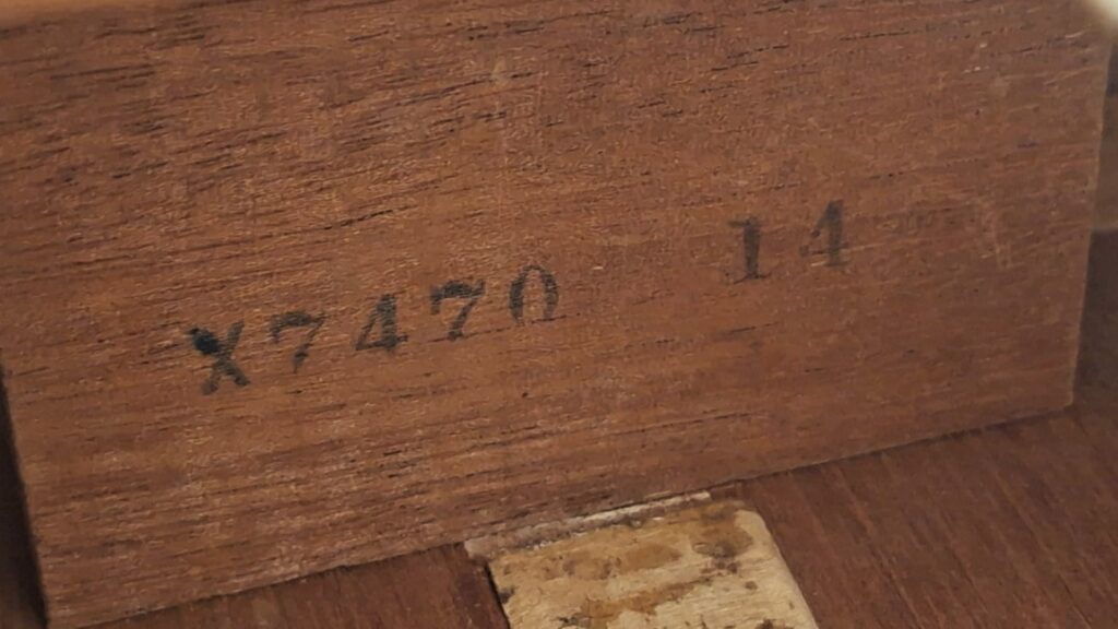 Nahaufnahme der Holzoberfläche einer Gibson LG-3 Akustikgitarre aus dem Jahr 1954, auf der der schwarze, eingeprägte Schriftzug „X7470 14“ sowie ein Teil eines abgenutzten Papieretiketts darunter zu sehen ist. Nahaufnahme der Holzoberfläche einer Gibson LG-3 Akustikgitarre aus dem Jahr 1954, auf der der schwarze, eingeprägte Schriftzug „X7470 14“ sowie ein Teil eines abgenutzten Papieretiketts darunter zu sehen ist.