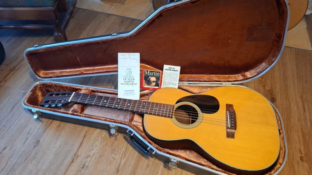 Eine Martin 00-21 Akustikgitarre aus dem Jahr 1986 liegt in einem offenen, gefütterten Hartschalenkoffer auf einem Holzboden. Im Koffer befinden sich neben dem Hals der Gitarre mehrere Broschüren zur Gitarrenpflege sowie eine Flasche Martin-Gitarrenpolitur. Eine Martin 00-21 Akustikgitarre aus dem Jahr 1986 liegt in einem offenen, gefütterten Hartschalenkoffer auf einem Holzboden. Im Koffer befinden sich neben dem Hals der Gitarre mehrere Broschüren zur Gitarrenpflege sowie eine Flasche Martin-Gitarrenpolitur.
